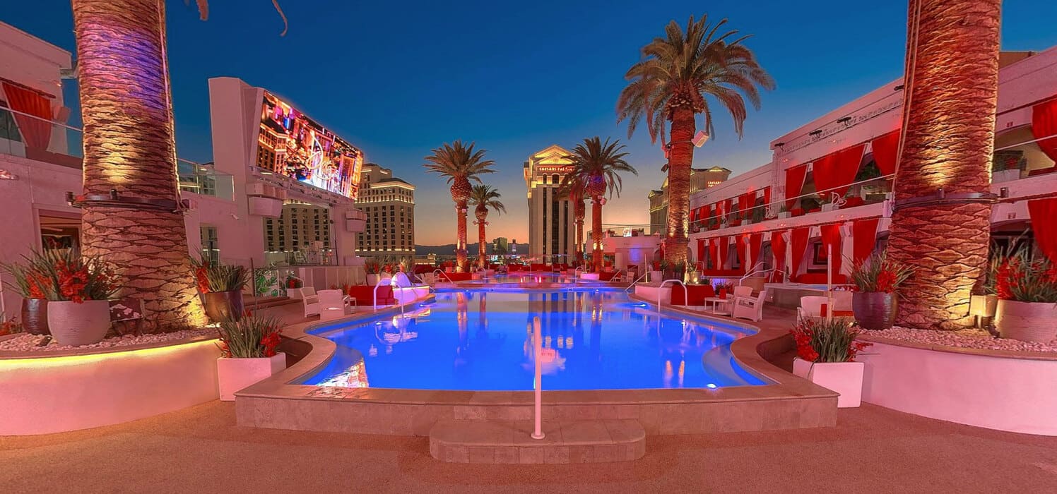 Rooftop pool at dusk with glowing blue water, palm trees, red cabanas, and city skyline views.